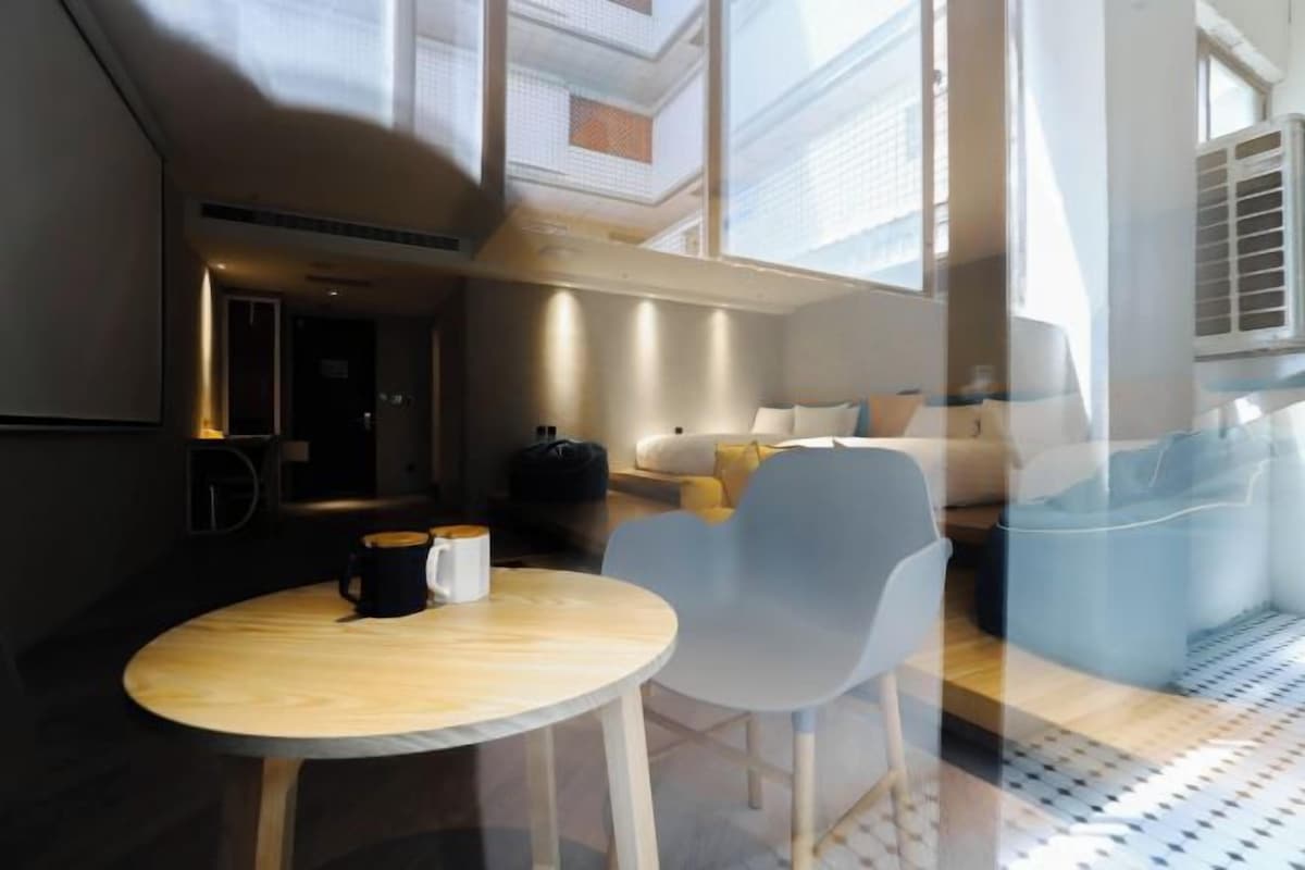 A modern hotel room with a round wooden table, two chairs, a sofa, and a large window providing natural light.