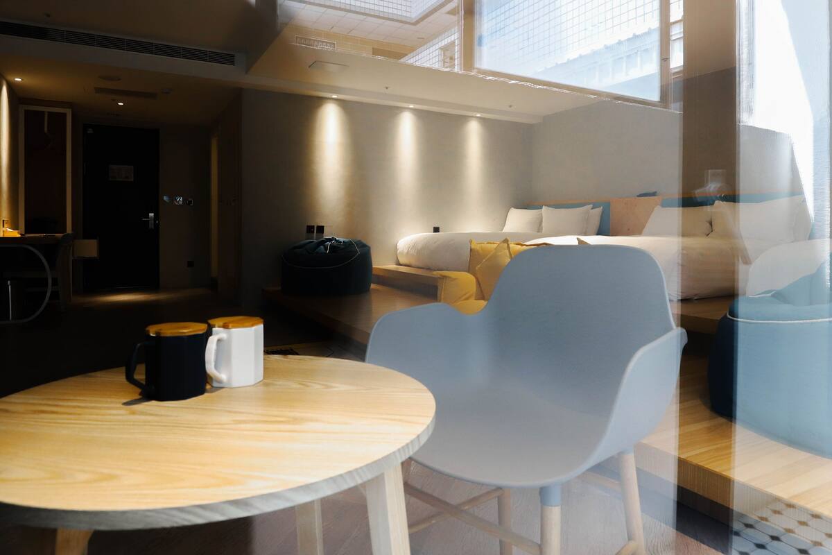 A modern hotel room with a bed, a seating area with a table and chairs, and a view through a window.