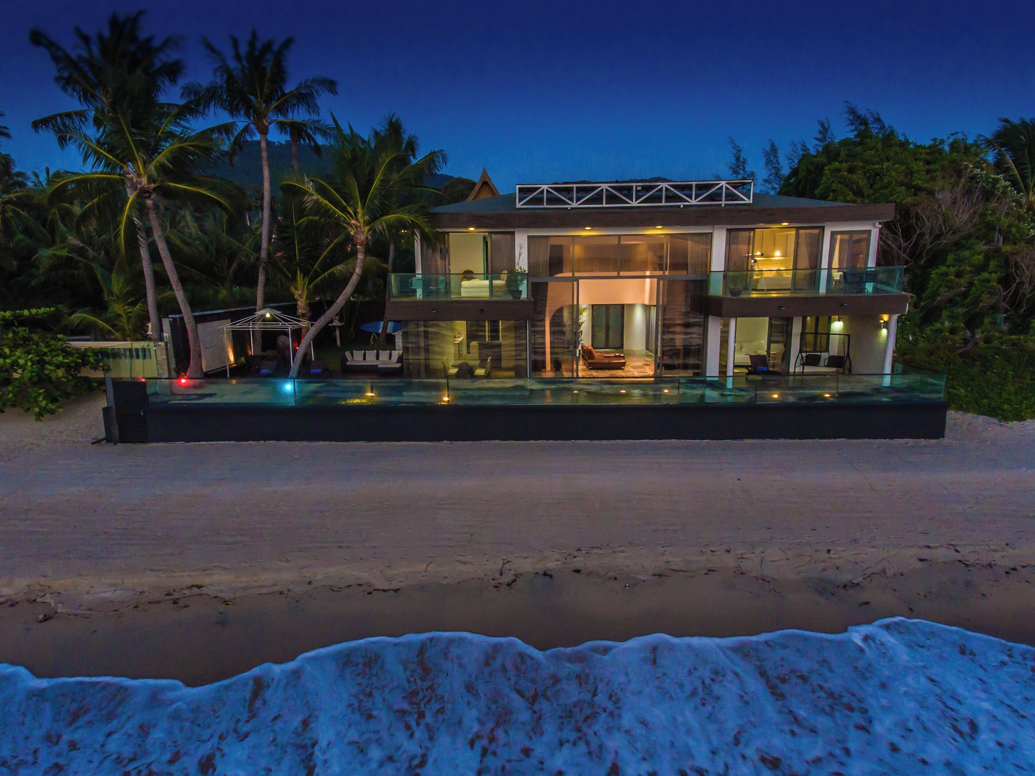 A beachfront property with a modern design, featuring a large glass facade, a pool, and a well-lit outdoor area.