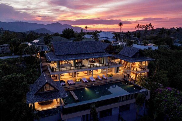 A multi-story resort with a swimming pool, surrounded by tropical vegetation and palm trees, during sunset.