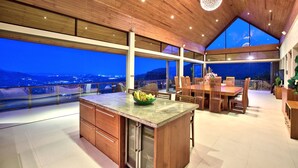A modern kitchen with a large island, wooden cabinetry, and a view of the city at dusk. - Grand Vista - 3 Bed, Sleep 6 (Ko Samui)