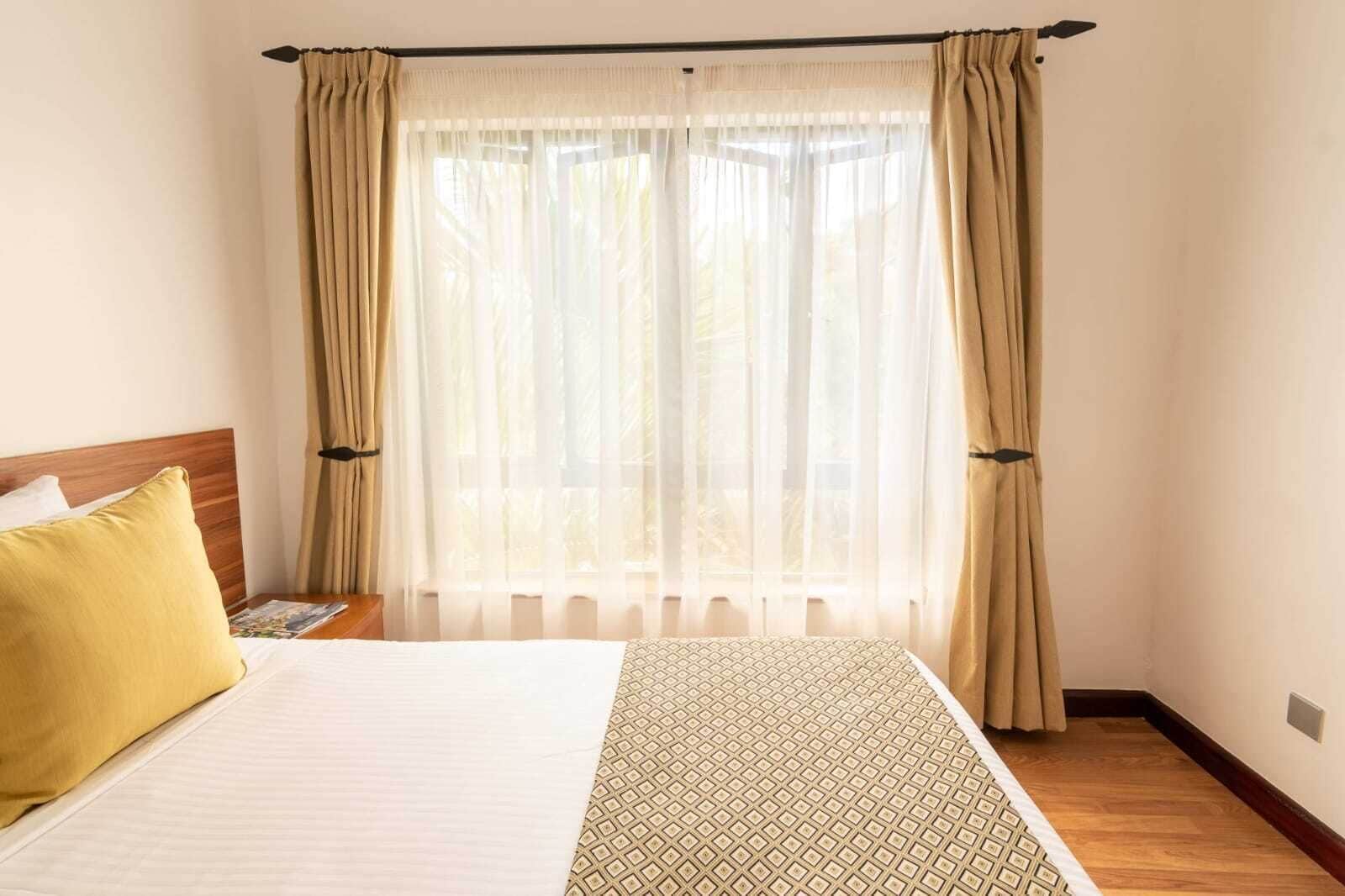 Ein Hotelzimmer mit einem Bett, einem Nachttisch, einem gelben Kopfkissen und einem Fenster mit durchsichtigen Vorhängen.