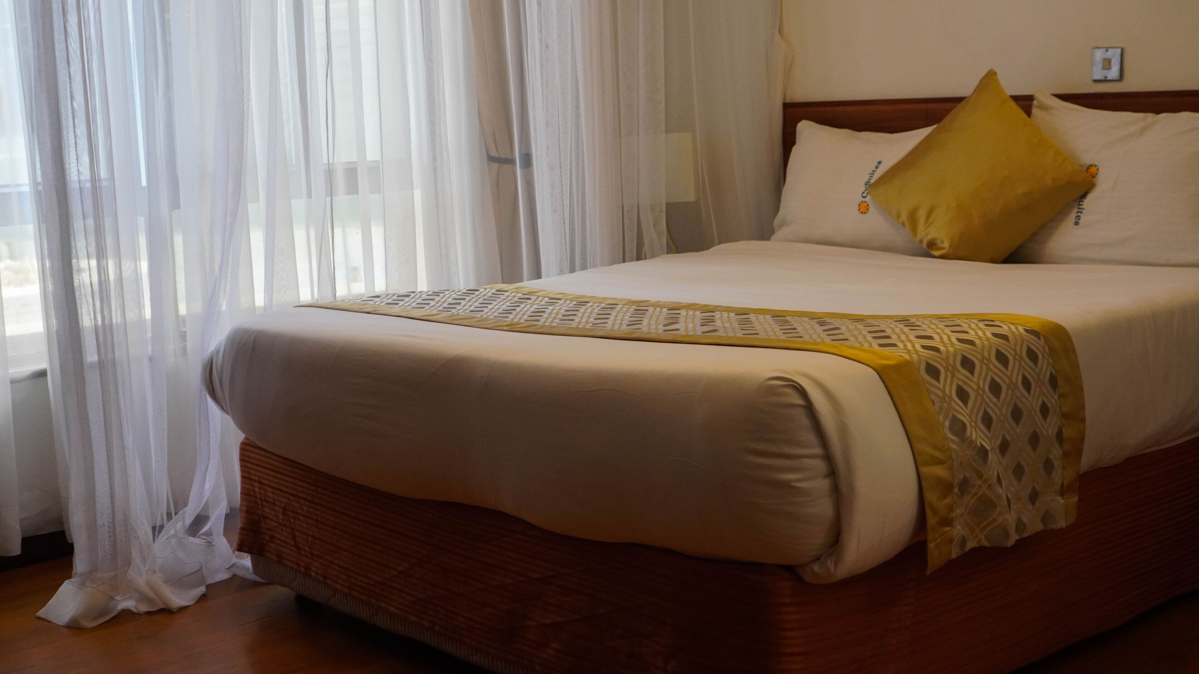 Una cama bien hecha con una colcha estampada y un corredor decorativo, una almohada amarilla y cortinas translúcidas corridas a un lado.