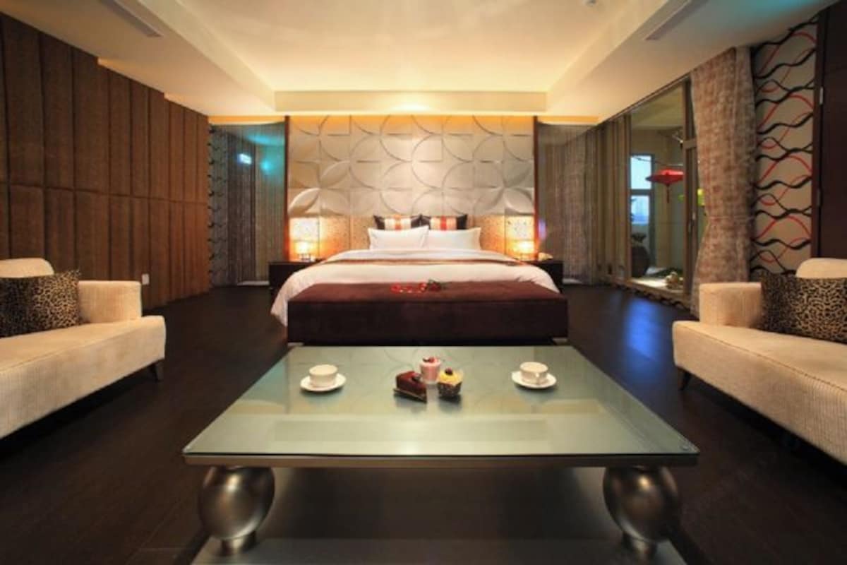 Una habitación de hotel moderna con una cama grande, dos sofás, una mesa de centro de cristal y pasteles, además de un cabecero decorativo.