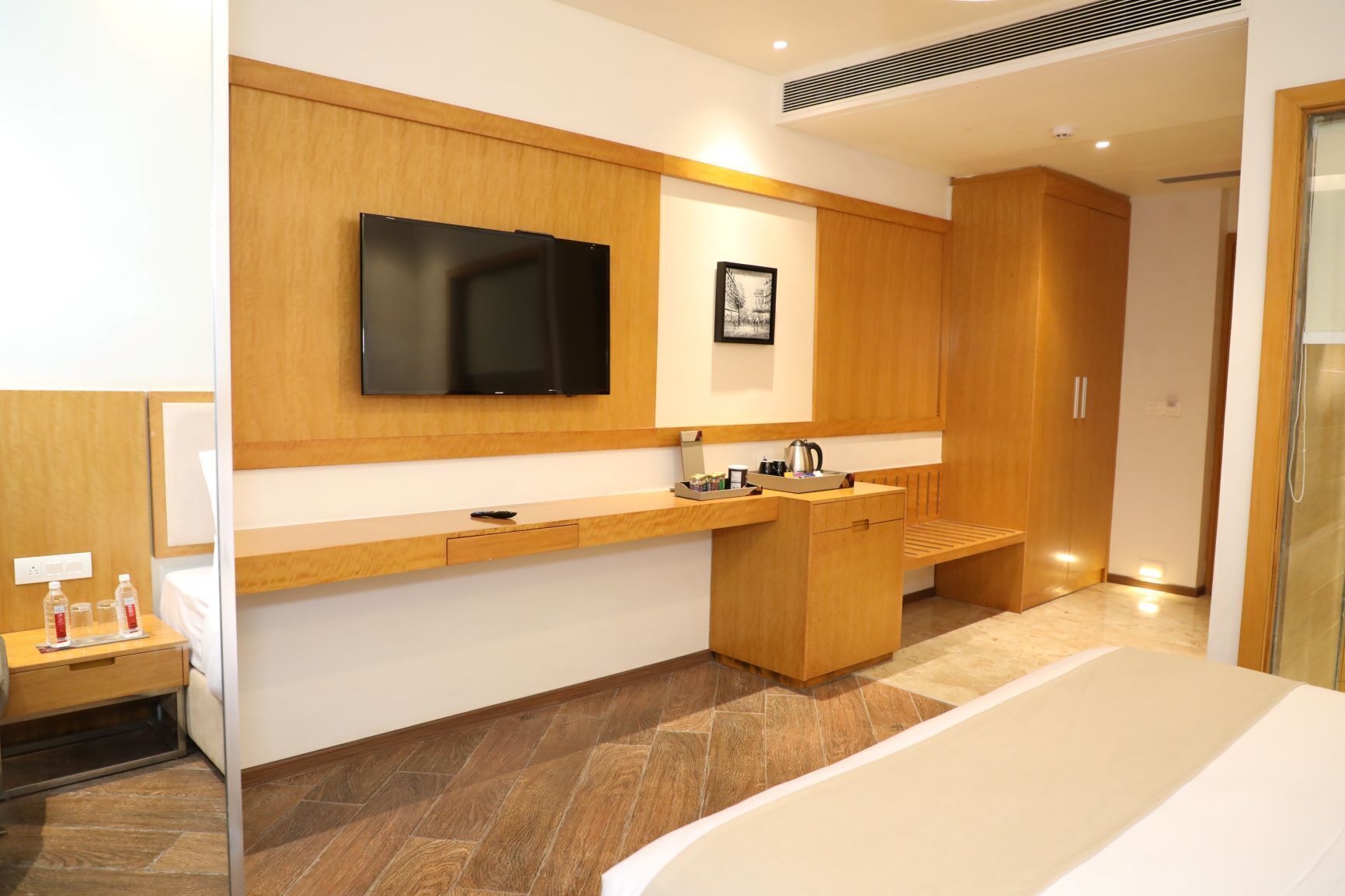 Una habitación de hotel moderna con un tocador de madera, un televisor de pantalla plana montado en la pared, una mesita de noche con artículos de aseo y una cama grande.