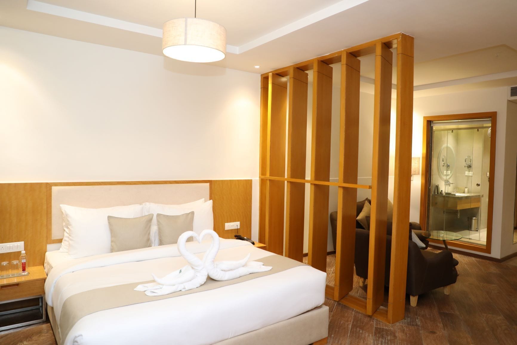 Habitación de hotel con una cama grande, un separador de madera, cabina de ducha con vidrio y una mesita de noche con lámpara.
