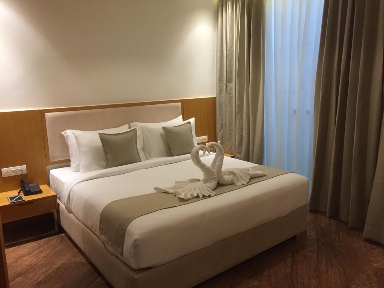 A hotel room with a neatly made bed, a bedside table with a phone and a notepad, and a window with curtains.