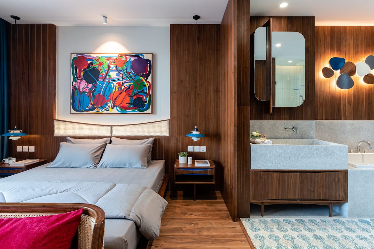A modern hotel room with a large bed, a wooden headboard, a colorful abstract painting, a built-in mirror, and a bathroom with a large sink and a bathtub.