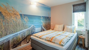 A bedroom with a mural of a beach scene, a bed with striped bedding, and a window with a view of greenery. - Oud Kempen Bungalow 99 (Stavenisse)