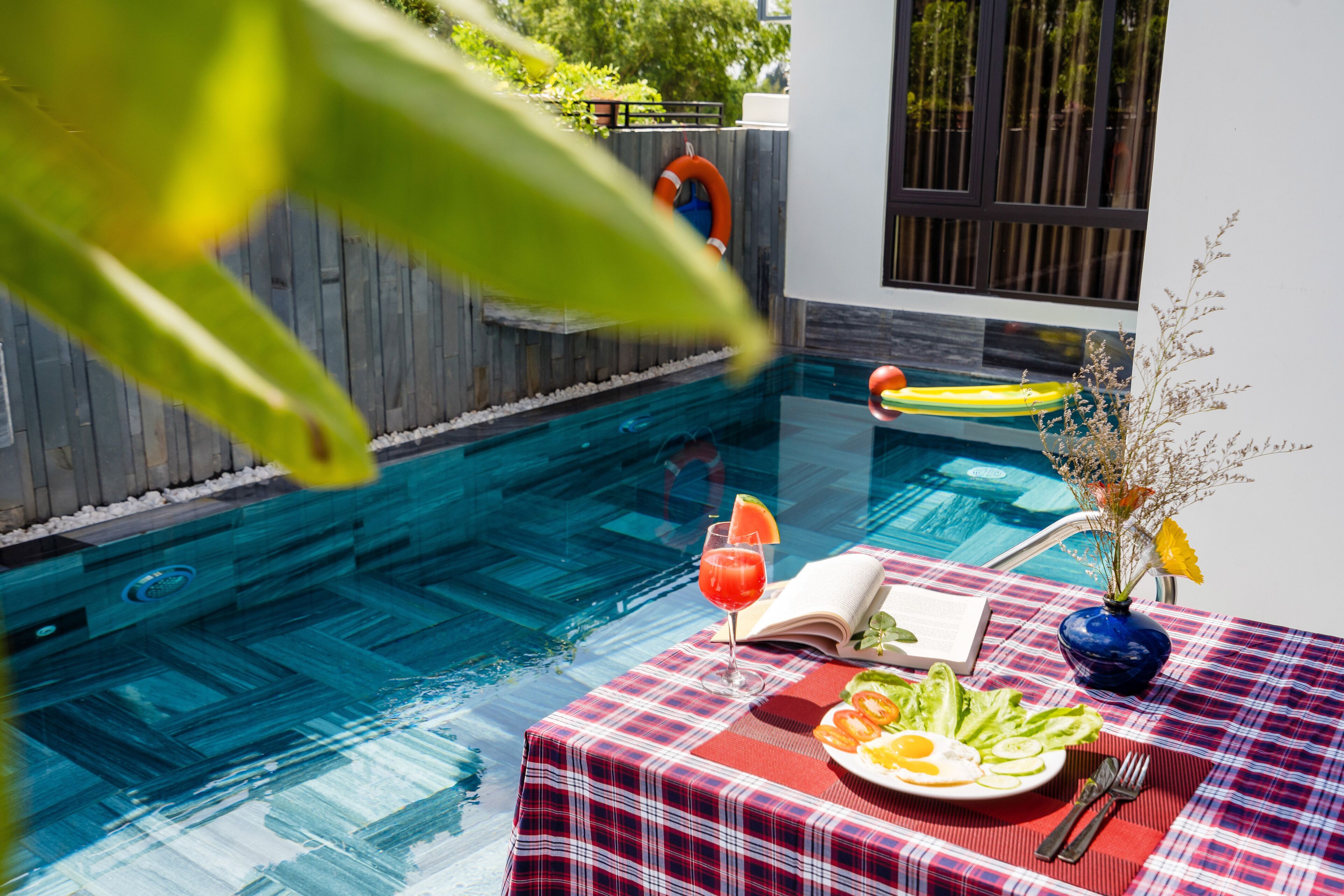 A poolside table with a red and white checkered tablecloth, a plate of food, a drink, and a book.