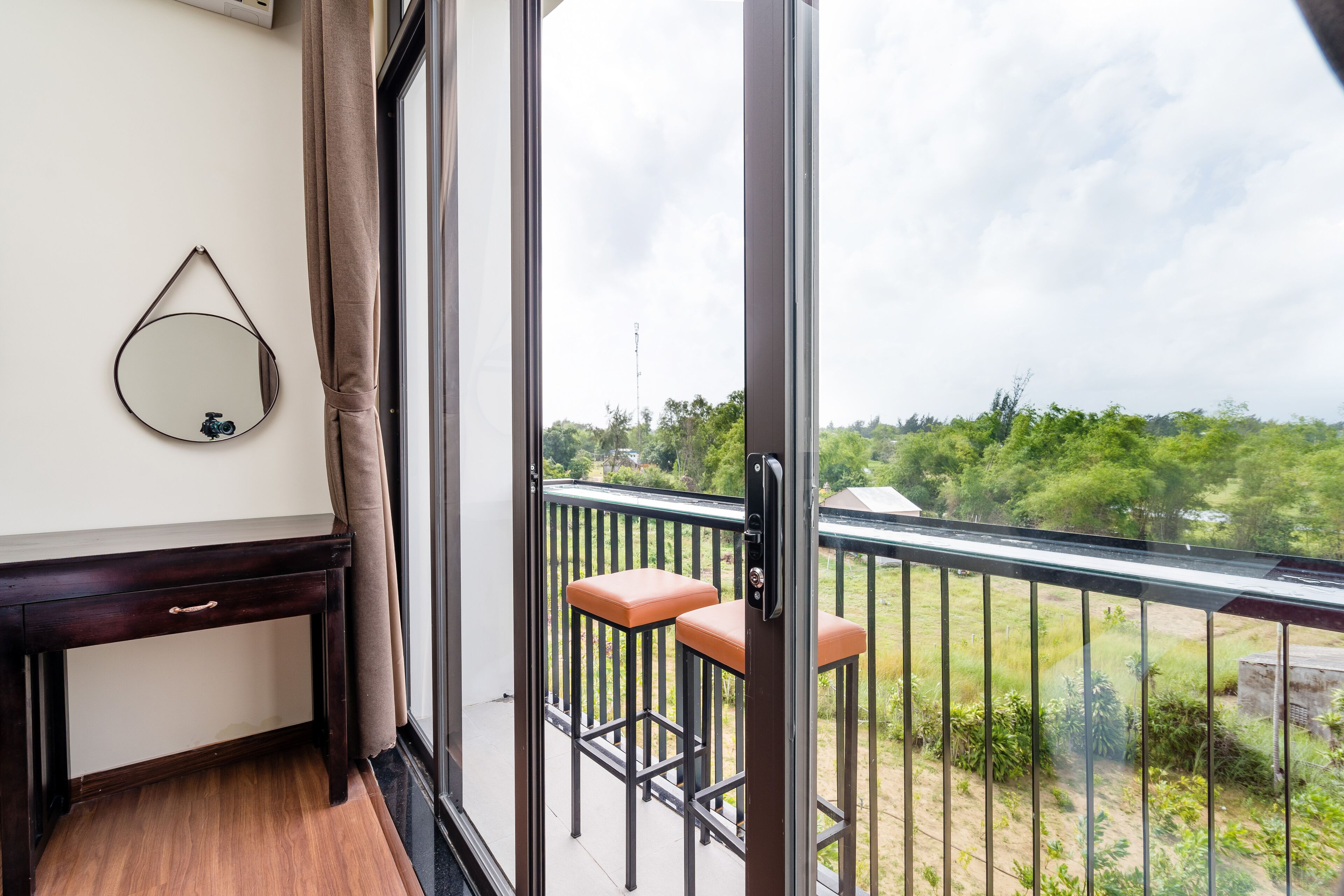 A balcony with a view of greenery, a round mirror, and a wooden desk with a drawer.