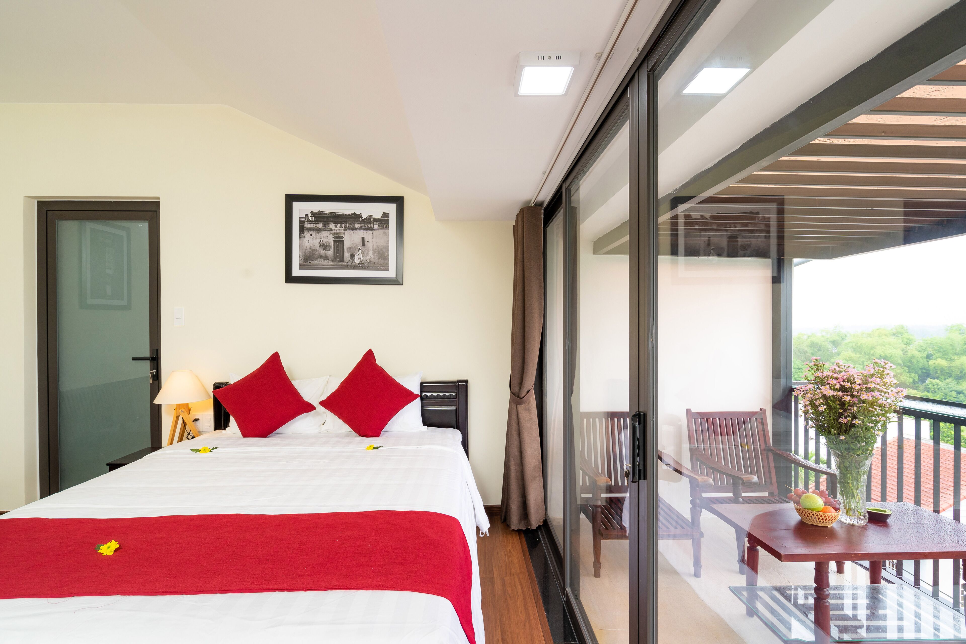 A hotel room with a bed, a nightstand, a lamp, a chair, a table with a vase, and a balcony with a view.