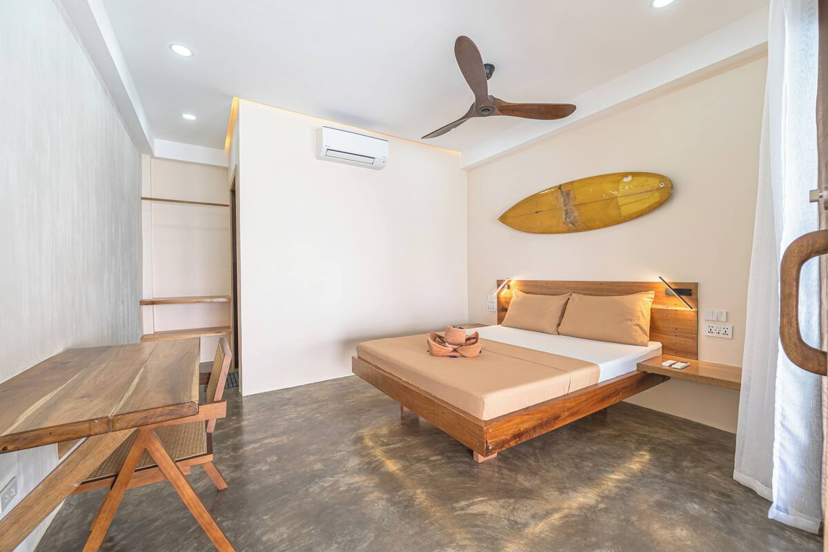 A bedroom with a bed, a ceiling fan, a wall-mounted air conditioner, a wooden table, and a surfboard decoration.
