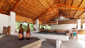 A pool table with two bottles of Red Horse beer, a wooden table, and a thatched roof structure. - Sunlit Hostel (General Luna)