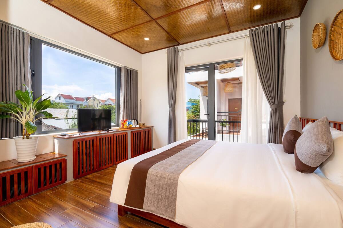 Ein geräumiges Schlafzimmer mit einem großen Bett, Holzboden, einem Fernseher auf einer Holzkonsole und einem Balkon mit Blick auf die Stadt.