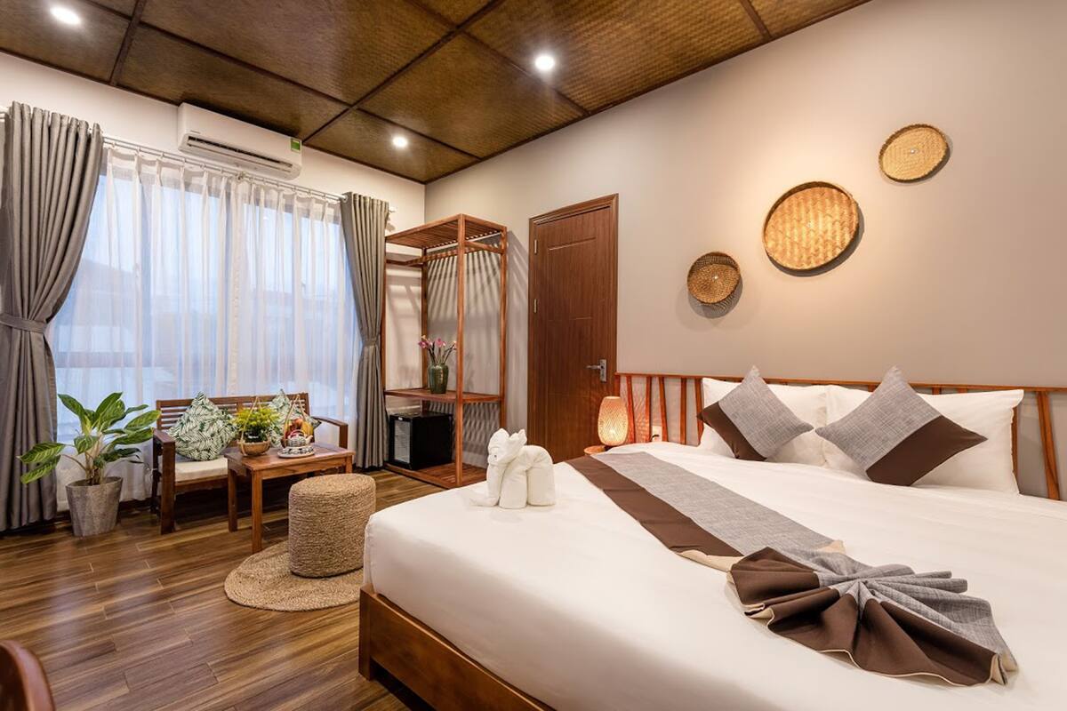 Ein Hotelzimmer mit Bett, Holz-Kopfteil, Nachttisch mit Lampe, Stuhl, kleinem Tisch mit Pflanze und Kleiderschrank.