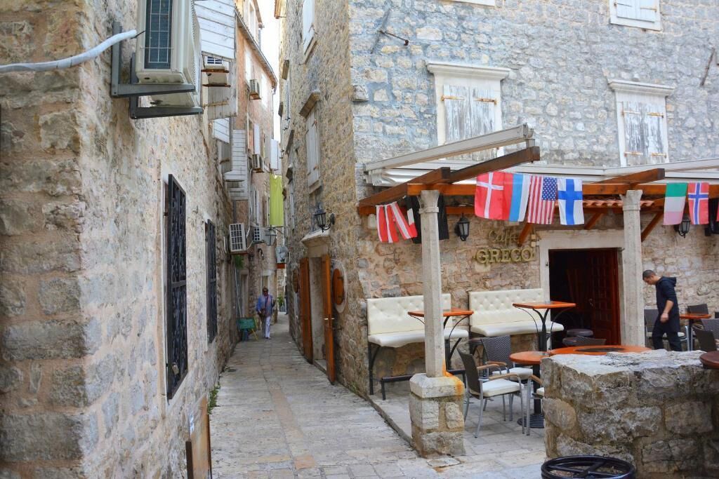 A narrow street with stone buildings, outdoor seating, and flags.