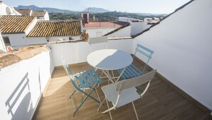 A rooftop terrace with a white table and blue chairs, overlooking a cityscape with terracotta roofs and distant mountains.