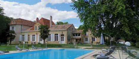 Una piscina con sillones de descanso, una casa grande con techo de tejas y un árbol al fondo.