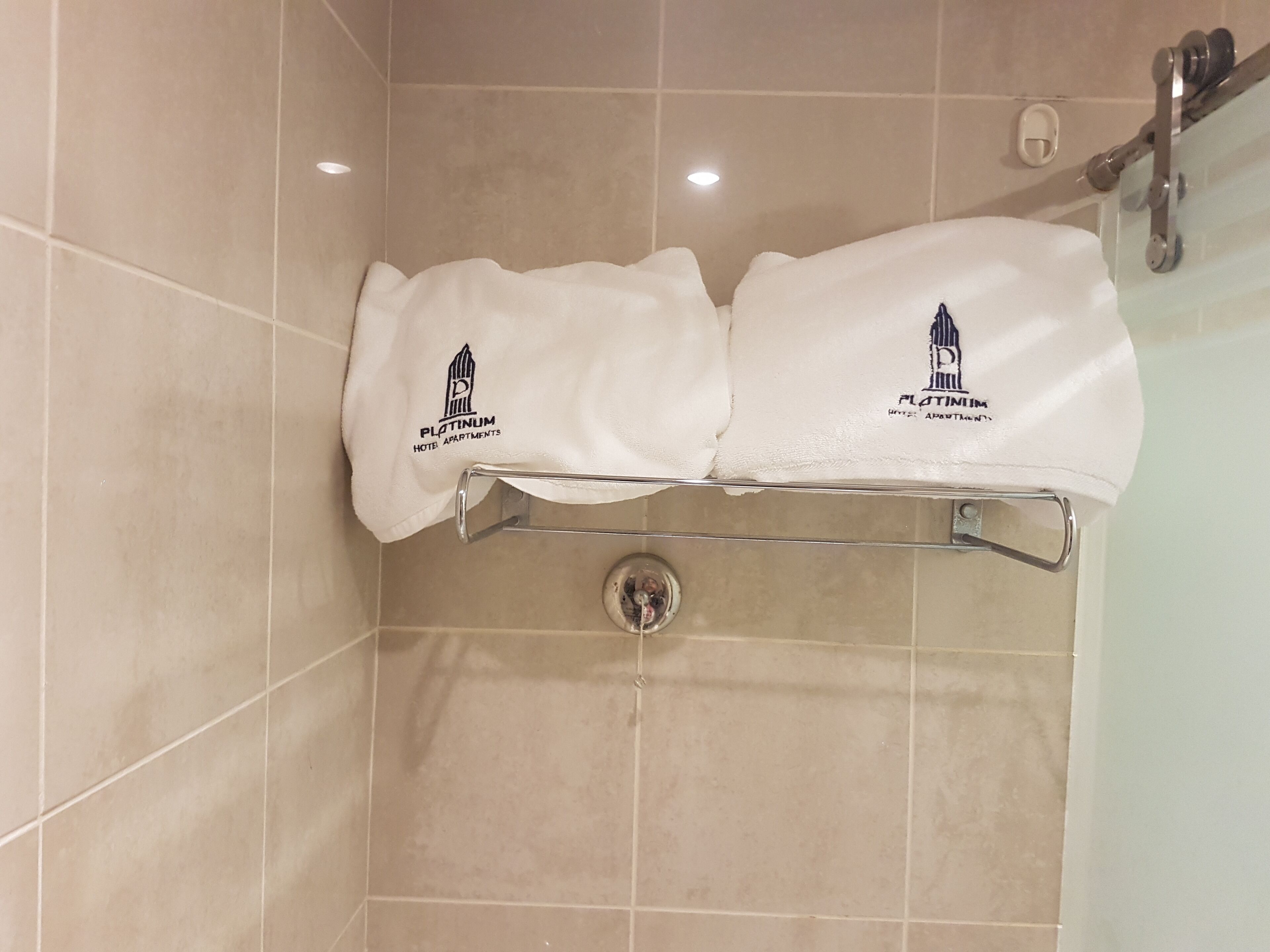 Una zona de ducha con azulejos color beige, una puerta de cristal y dos toallas blancas colgadas en un soporte metálico con logotipos.