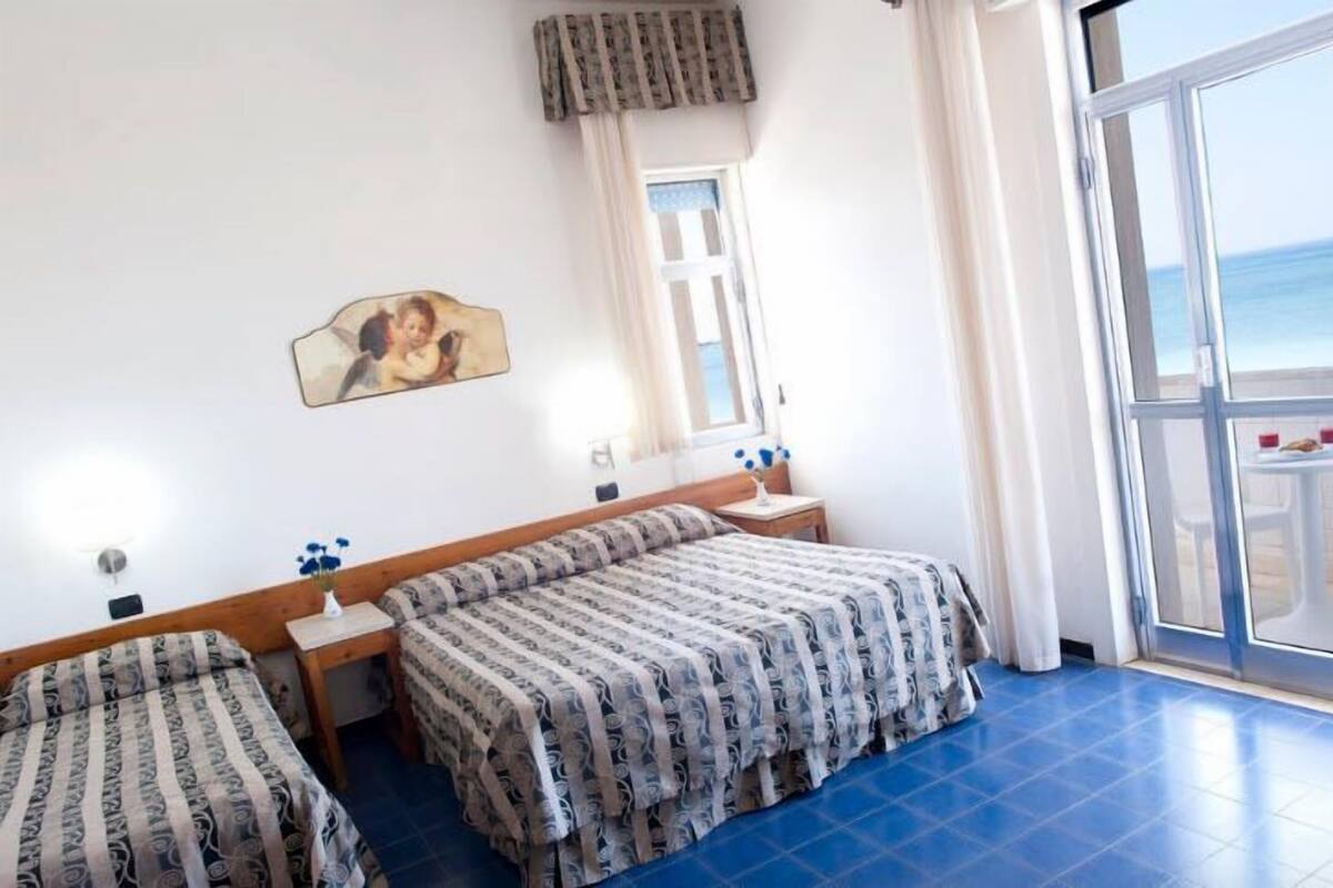 Una habitació d'hotel amb dos llits, una finestra amb vista al mar i un balcó amb taula i cadires.