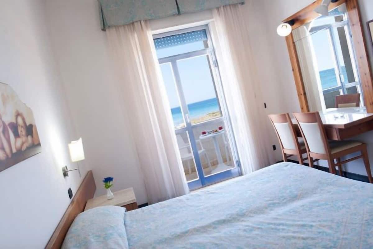 Habitació d'hotel amb un llit, una taula de treball amb cadires, una finestra que dona al mar i un quadre a la paret.