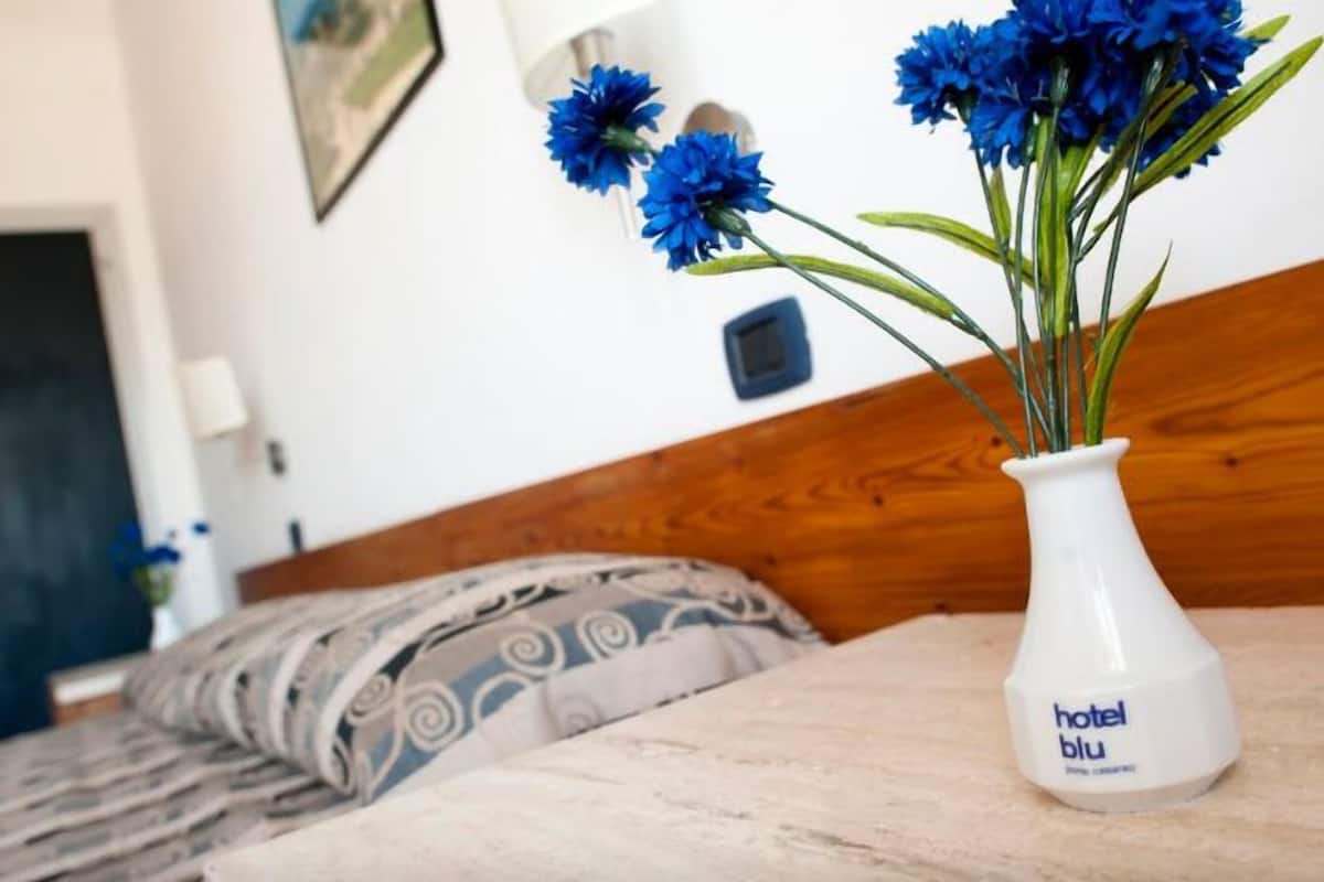 Habitació d'hotel amb un llit, un ram de flors blaves en un vas blanc i un panell de llit de fusta.