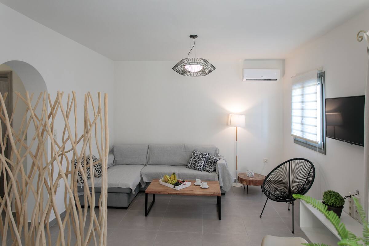 A modern living room with a grey sofa, a wooden coffee table with fruits, a black chair, and a hanging light fixture.