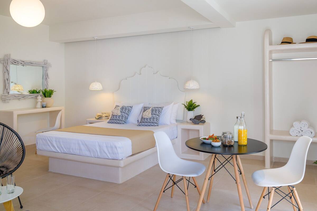 A modern bedroom with a bed, bedside tables, a round table with chairs, a wardrobe, and a mirror.