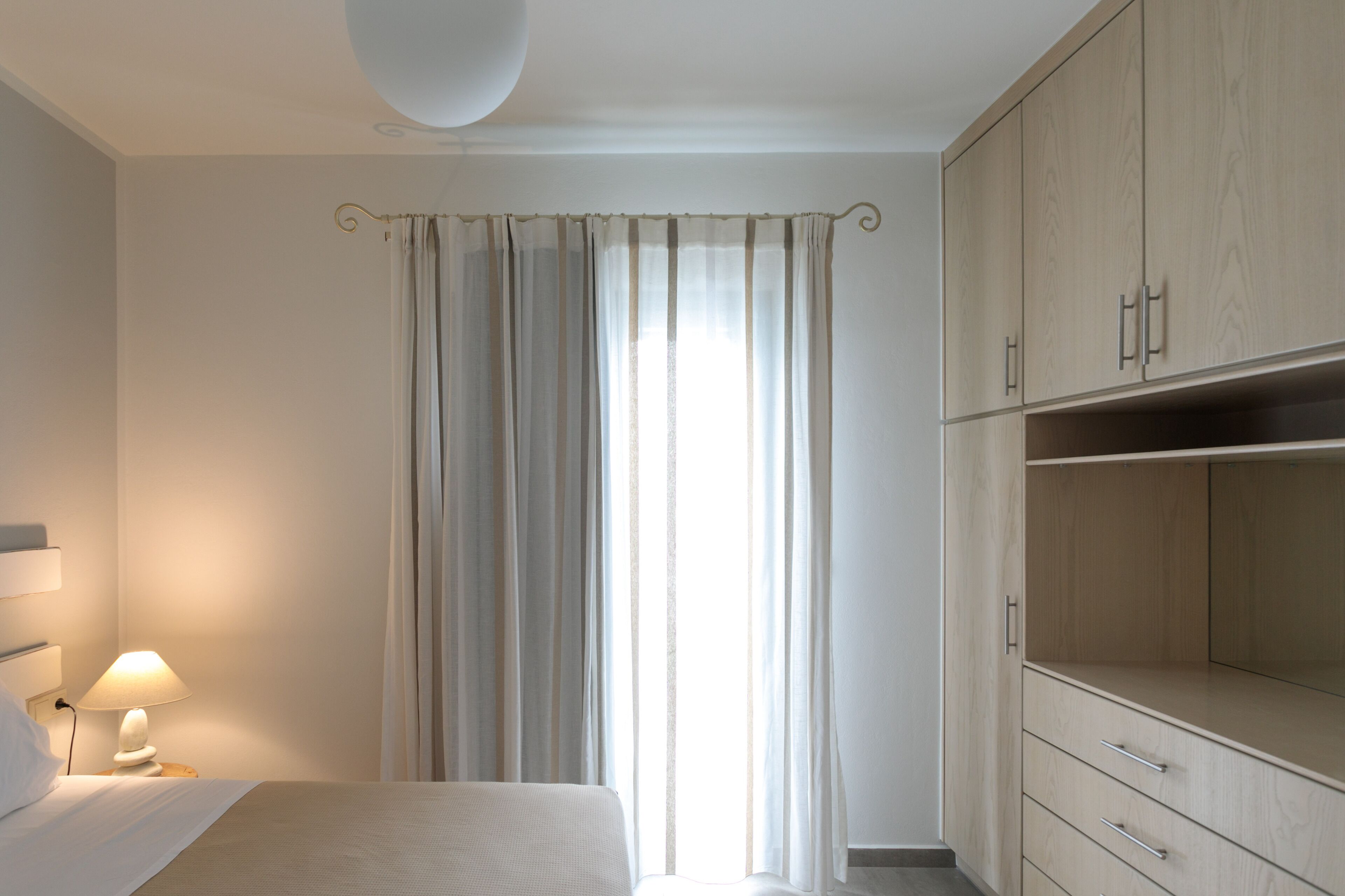 Une chambre à coucher comprenant un lit, une table de chevet avec une lampe, une fenêtre avec des rideaux et des armoires encastrées en bois.