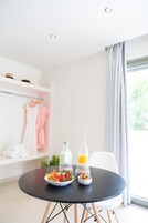 Una habitación moderna con una mesa de comedor redonda, un tazón de cereal con frutas, una botella de jugo de naranja y un vaso de leche. Hay una silla blanca y un estante con un vestido rosa y un sombrero.