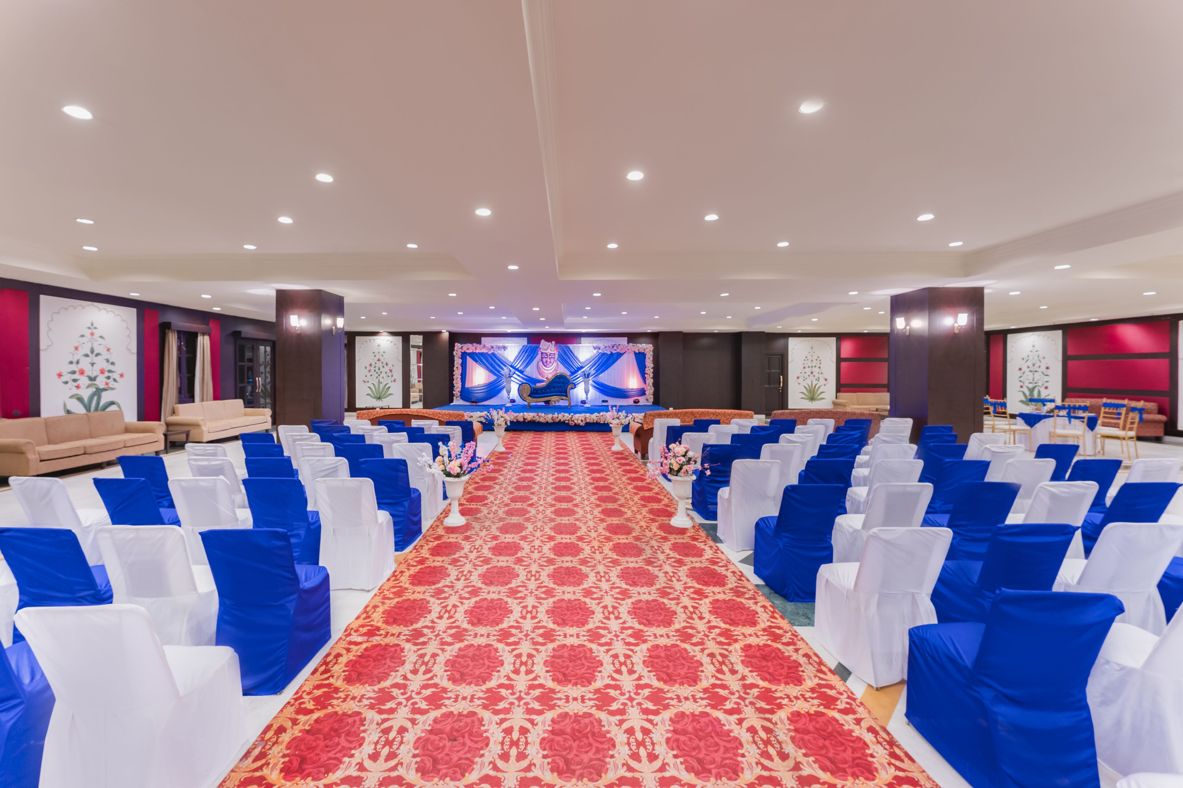 Un salón de banquetes con filas de sillas blancas y azules, una alfombra roja y un escenario con un telón de fondo azul.