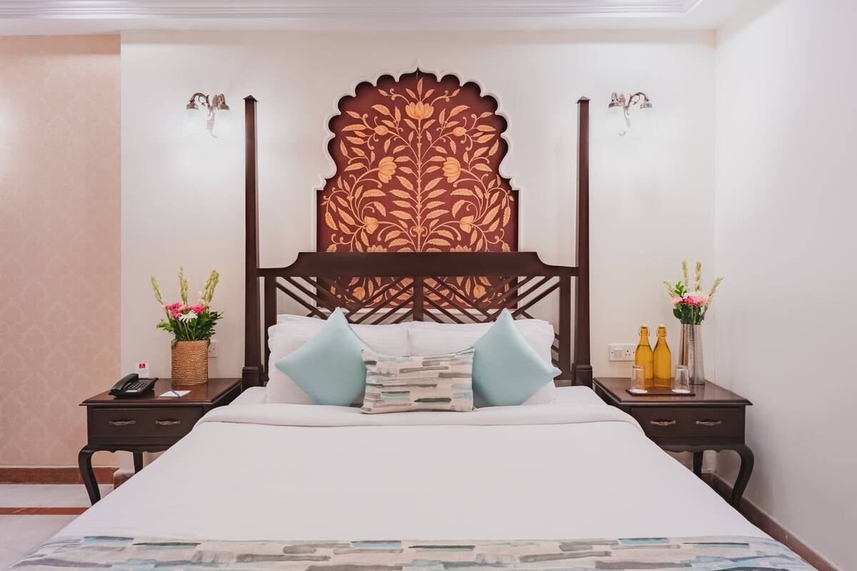 A hotel room with a large bed, a decorative headboard, two bedside tables with lamps, and a nightstand with a phone and a basket of flowers.