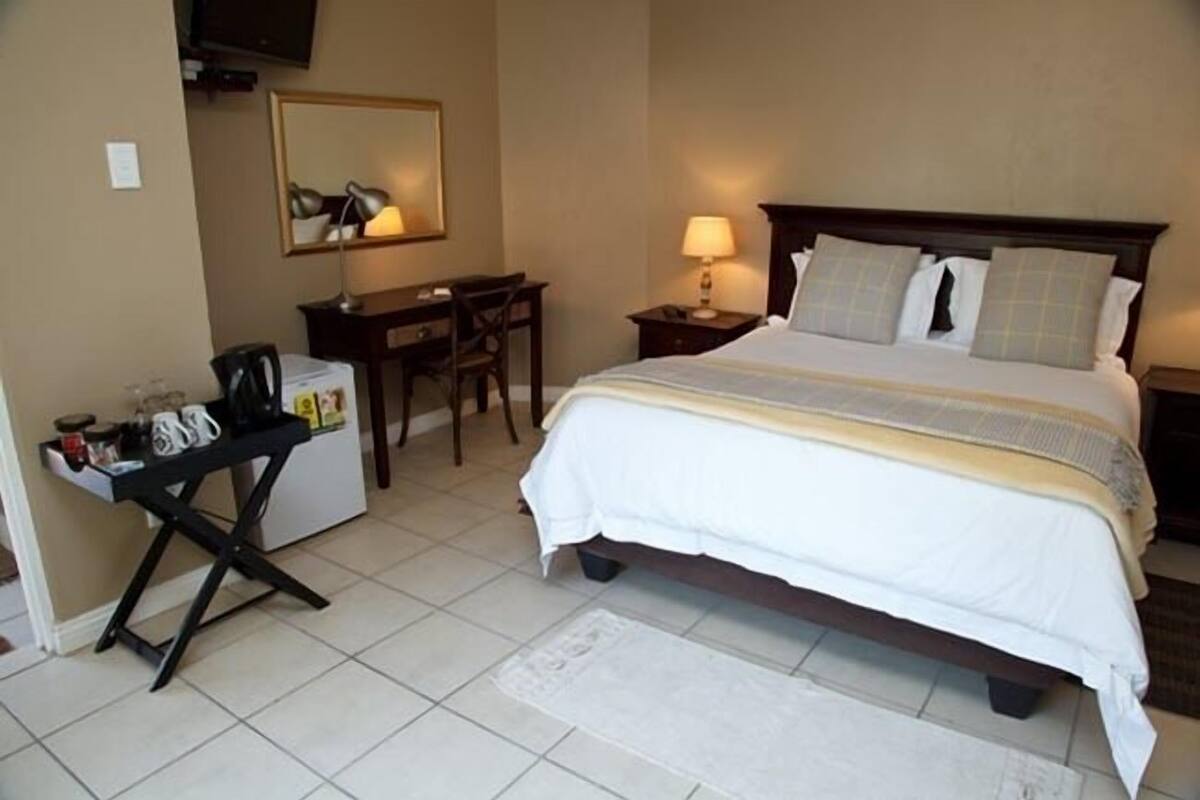 A hotel room with a bed, a desk, a chair, a lamp, and a small table with items on it.