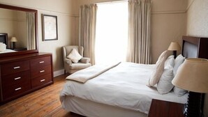 Habitación de hotel con una cama grande, un aparador de madera, una silla, un escritorio con lámpara y un amplio ventanal con cortinas.