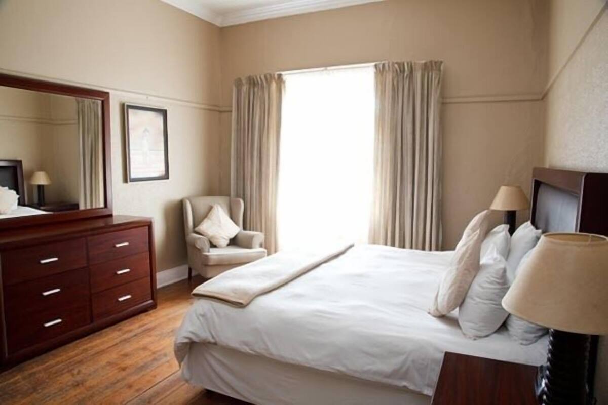 A hotel room with a large bed, a wooden dresser, a chair, a desk with a lamp, and a large window with curtains.