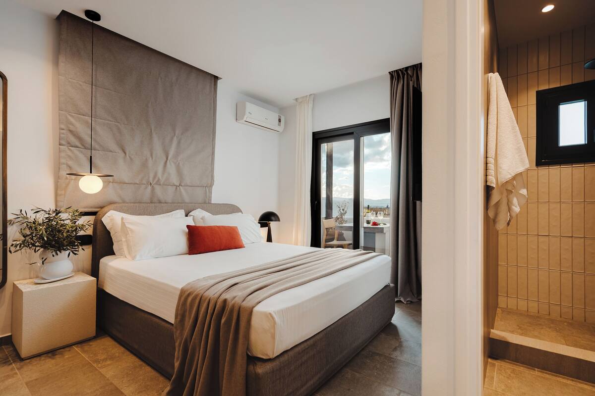 A modern hotel room with a large bed, a bedside table with a lamp, a potted plant, and a view of the outdoors.