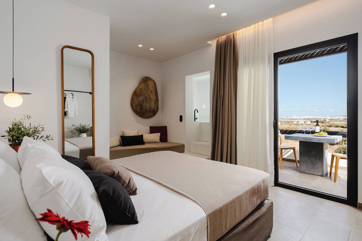 A modern bedroom with a large bed, a mirror, a view of the outdoors, and a balcony with a table and chairs.
