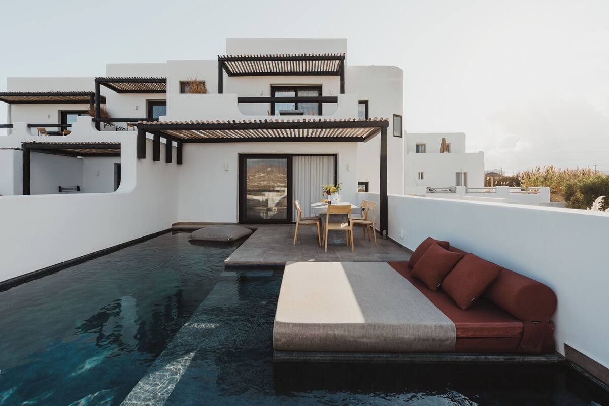 A modern building with a rooftop pool, a seating area with a sofa and table, and a covered patio with chairs.