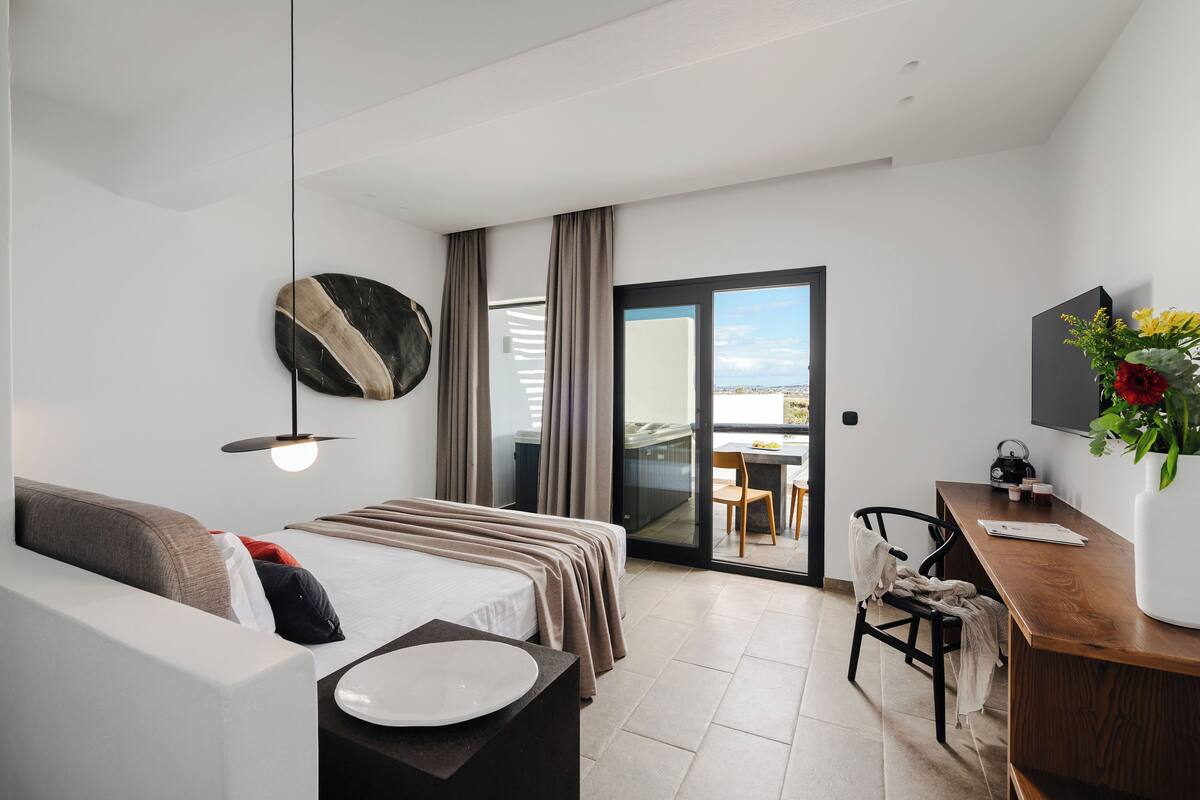 A modern hotel room with a bed, a desk, a chair, a TV, and a balcony with a view.