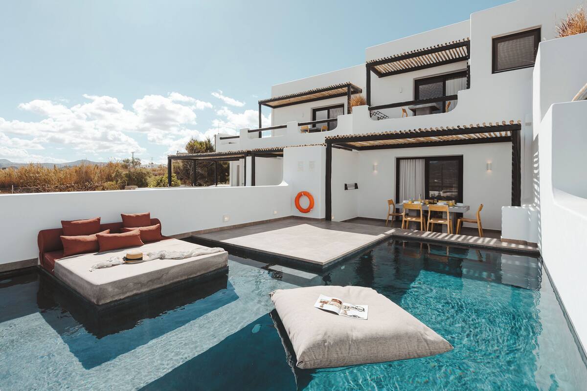 A modern outdoor pool area with a sofa and cushions, a dining table with chairs, and a clear view of the surrounding landscape.