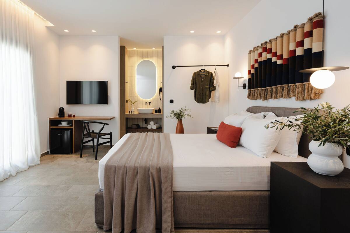 A modern hotel room with a large bed, a desk, a television, a mirror, and a wall-mounted artwork.
