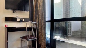 Una habitación con televisión en la pared, una silla, un ventanal con vista a un edificio y una cortina.