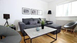 Una sala de estar moderna con un sofá gris, una mesa de centro y un sillón gris. Hay tres cuadros enmarcados en la pared, una lámpara de pie y un radiador.