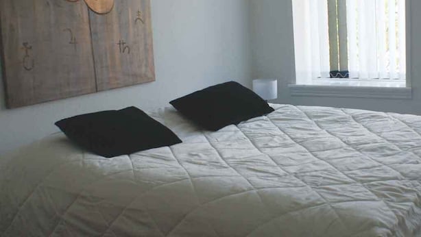 Una cama con colcha blanca y dos almohadas negras, una obra de arte de madera en la pared, y una ventana con cortinas.