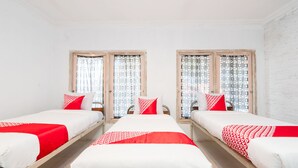 Une chambre d’hôtel avec deux lits simples, chacun recouvert d’une couvre-lit et agrémenté d’oreillers aux motifs rouges et blancs. Les murs de la chambre sont blancs, et un mur est décoré de briques blanches. Il y a deux fenêtres avec des rideaux à motifs, et le sol est carrelé.