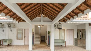 Un espace extérieur abrité, doté d’un plafond en bois, de murs blancs et d’un sol en briques. On y trouve deux chaises et une table, ainsi qu’un escalier menant à un niveau supérieur.