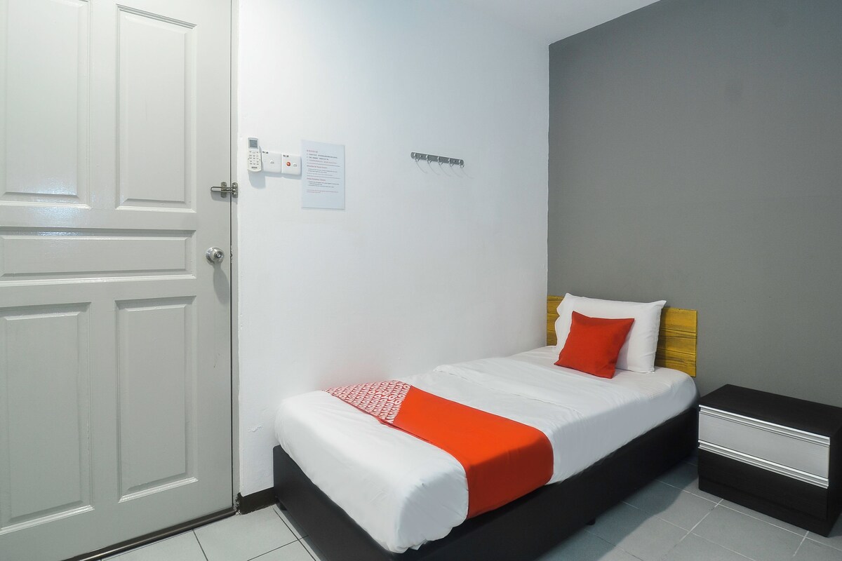 A single bed with white bedding and orange accents, a bedside table, a wall-mounted heater, and a door with a lock.