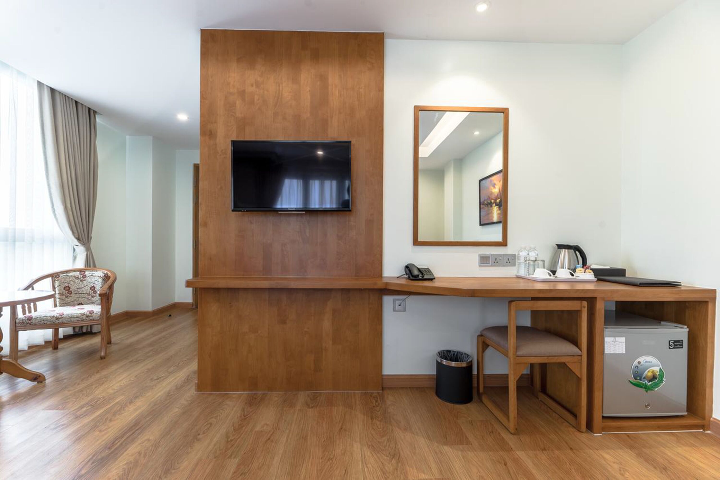 A hotel room with a wooden desk, a flat-screen TV mounted on a wooden panel, a mirror, a trash bin, and a small kitchenette area.