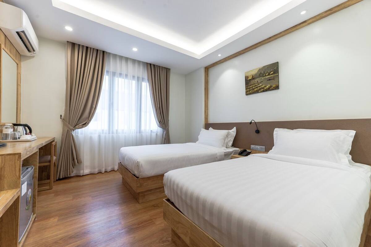 A hotel room with two beds, a desk with a coffee maker, a window with curtains, and a painting on the wall.